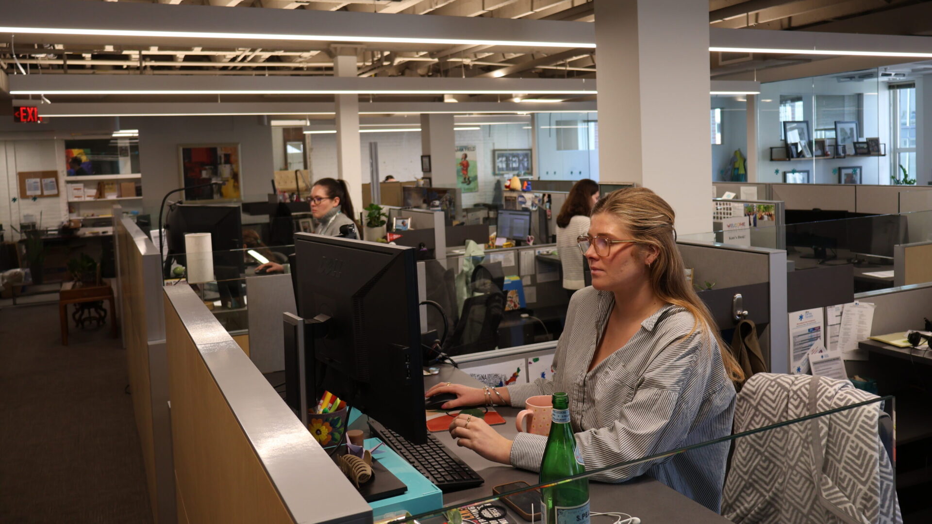 Office workspace with employees working at desks, using computers, surrounded by modern office furniture and open ceiling.