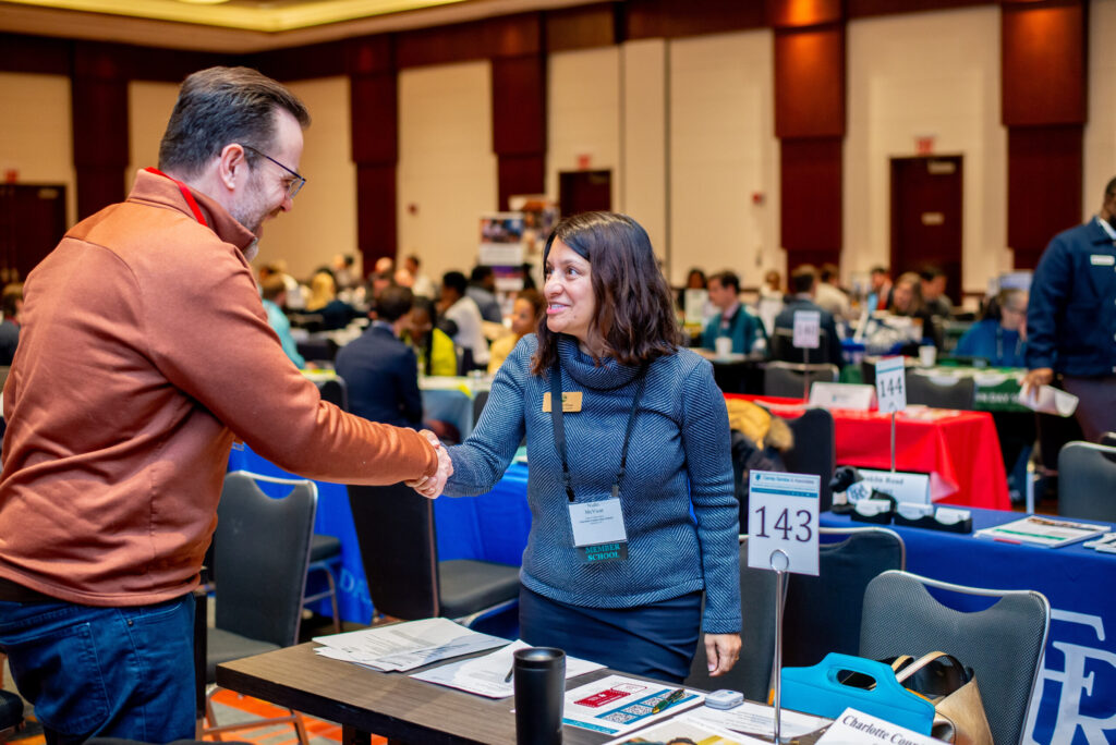 Two people shaking hands at a professional networking event, with tables and attendees in the background.