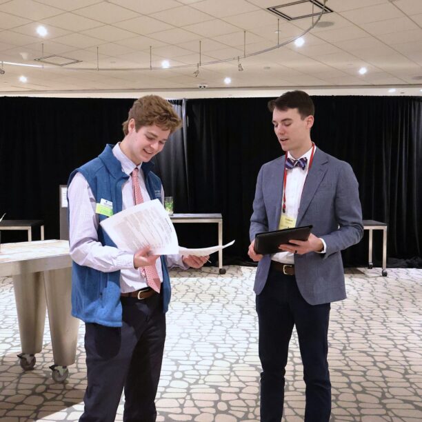 Two people standing with papers and a tablet in a conference room, discussing with tables and a curtain in the background.