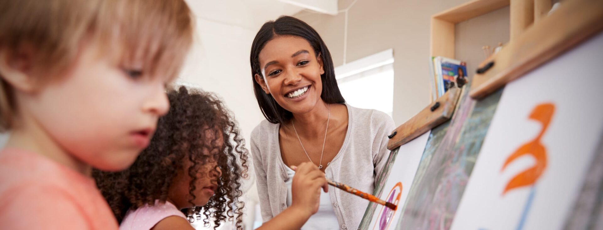 Children painting on easels with a smiling adult watching in a bright, cheerful classroom setting.