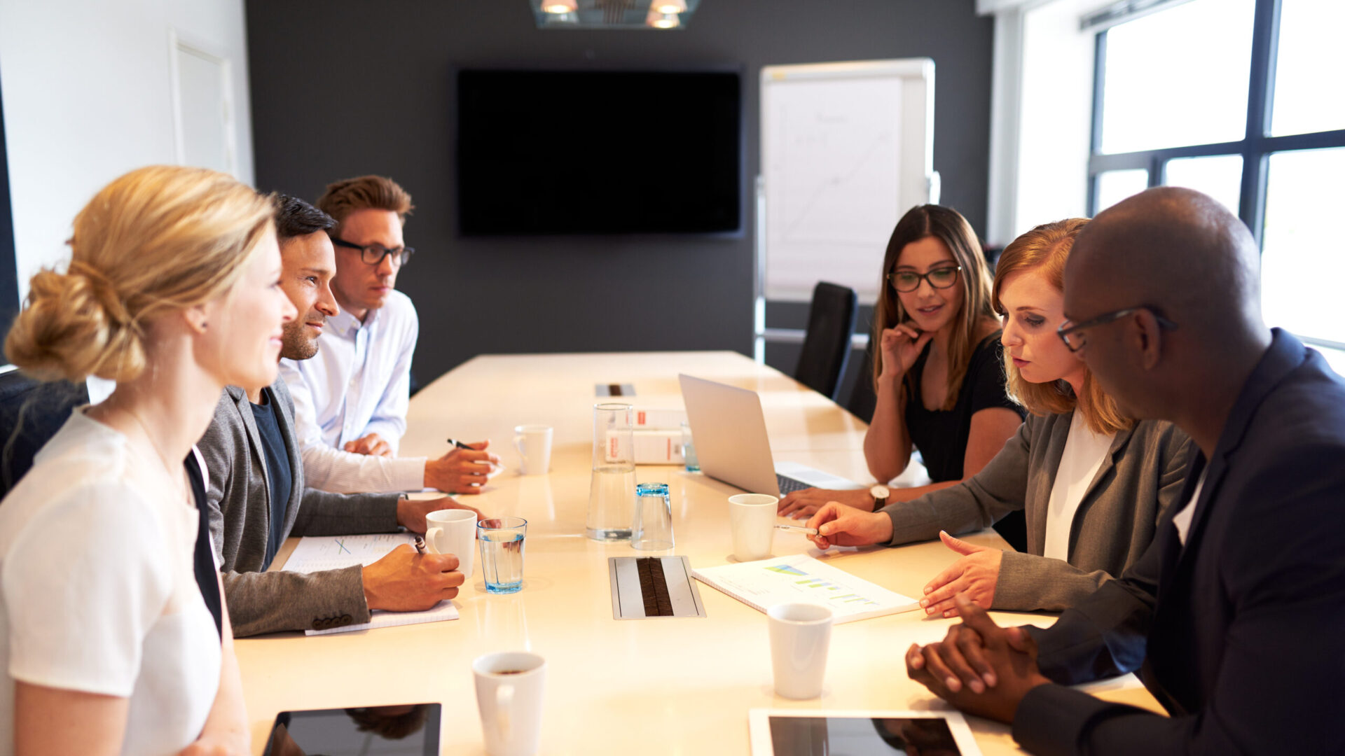 People in a business meeting around a long table with laptops and coffee cups in a modern office setting.