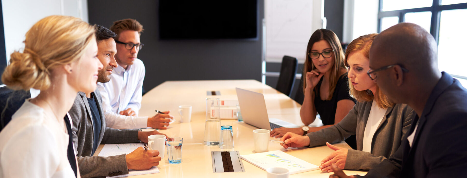 People in a business meeting around a long table with laptops and coffee cups in a modern office setting.
