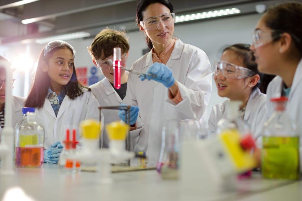 Science teacher demonstrating an experiment with students wearing lab coats and safety goggles in a classroom lab.