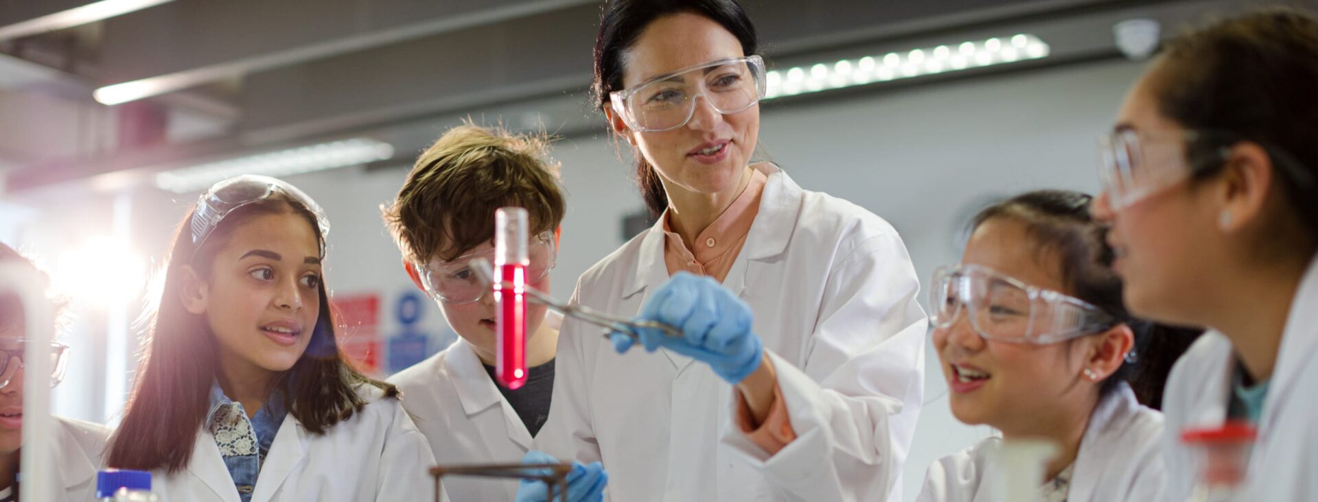Science teacher demonstrating an experiment with students wearing lab coats and safety goggles in a classroom lab.