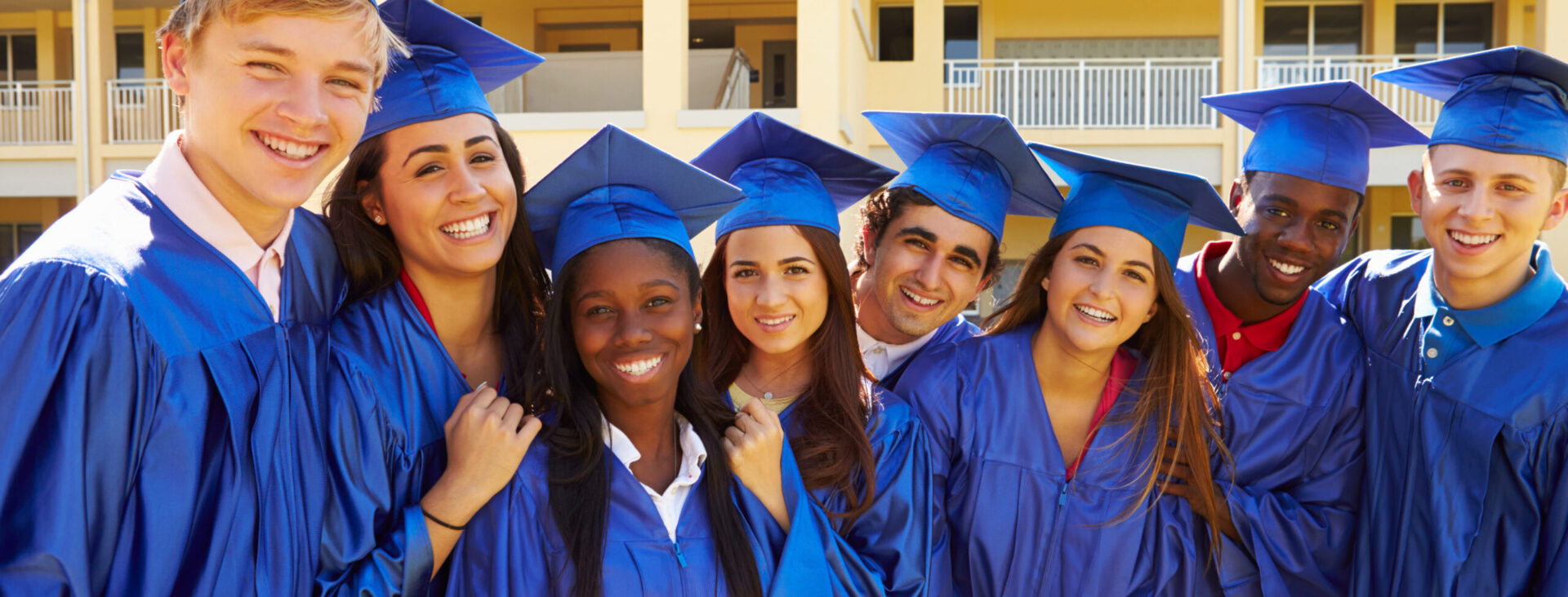 Group of happy graduates in blue gowns and caps standing together, smiling outdoors.