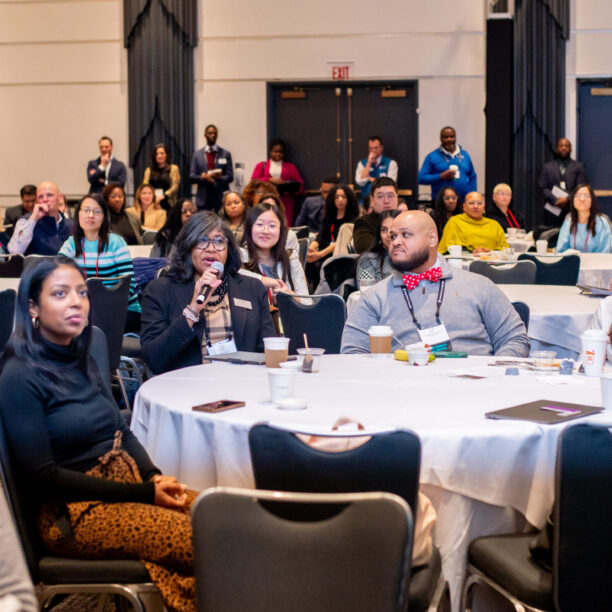 People sitting at round tables attending a conference, with some participants engaged and taking notes.