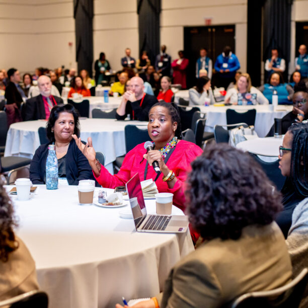 Audience engaged in a conference discussion at round tables with a woman speaking into a microphone.