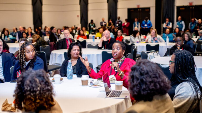 Audience engaged in a conference discussion at round tables with a woman speaking into a microphone.