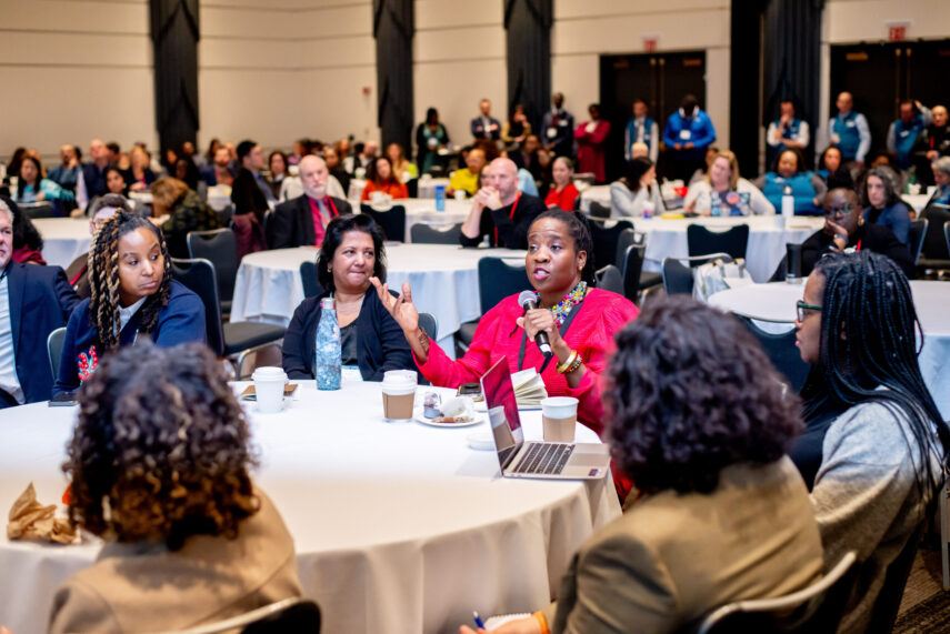 Audience engaged in a conference discussion at round tables with a woman speaking into a microphone.