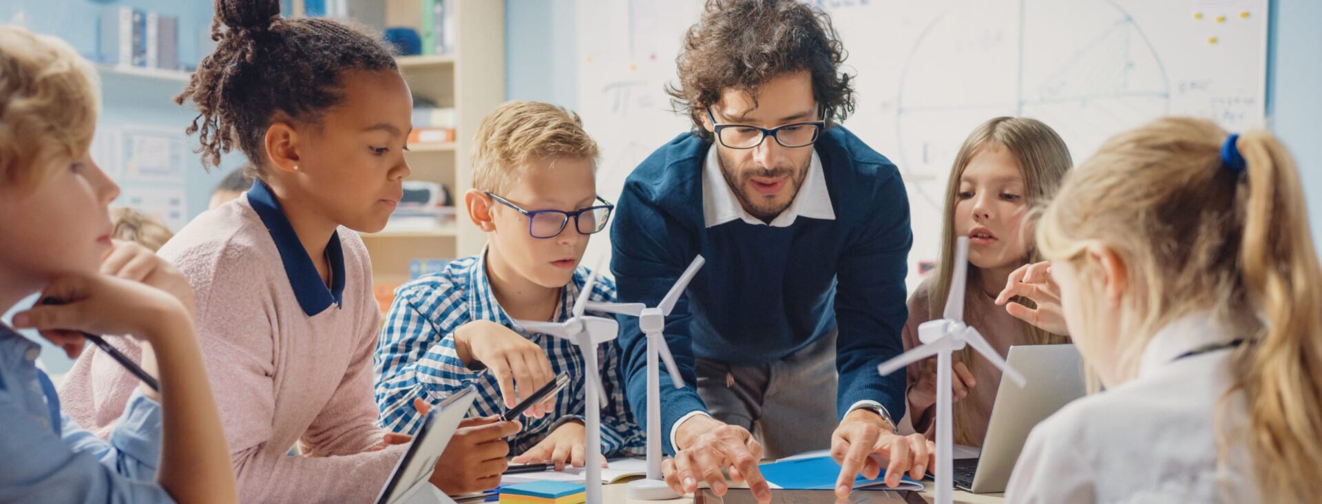 Teacher guiding diverse students in a classroom with wind turbine models and tablets on desks.