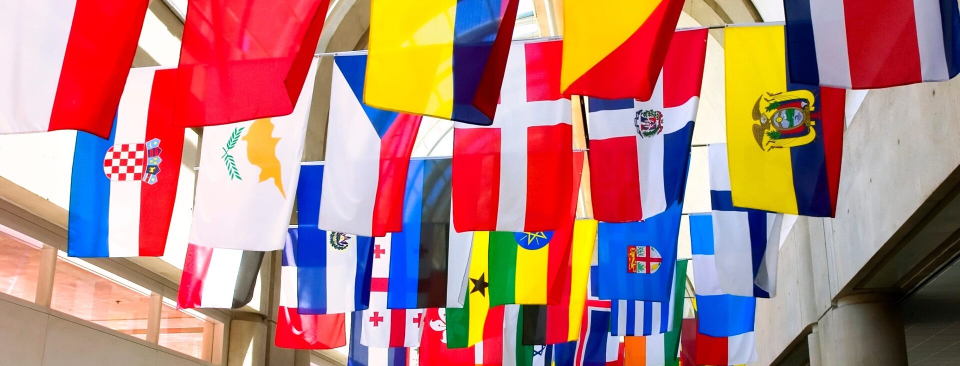 Colorful international flags hanging from a high ceiling in an indoor space with natural light filtering through windows