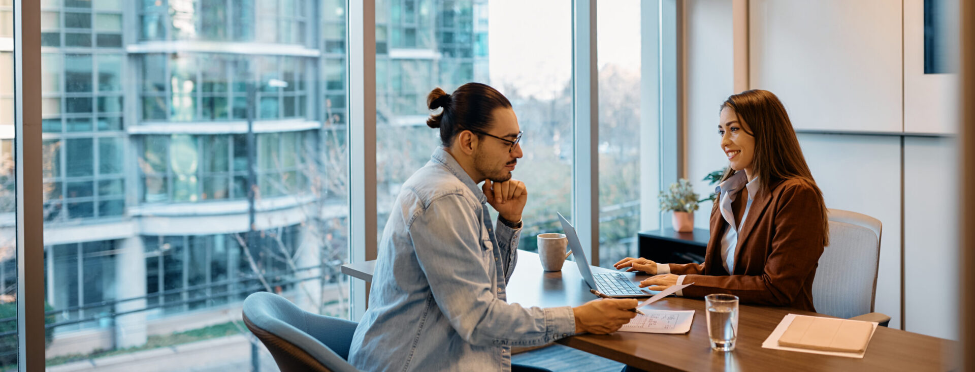 Two colleagues discussing work during a meeting in a modern office with large windows and a view of glass buildings outside.
