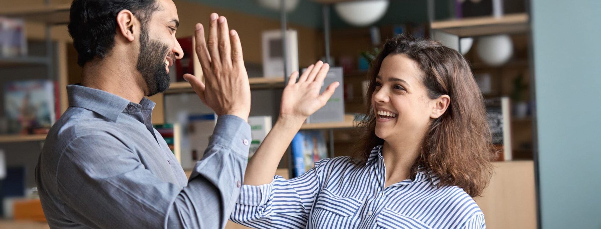 Two colleagues in an office giving each other a high-five, smiling, with shelves and books in the background.