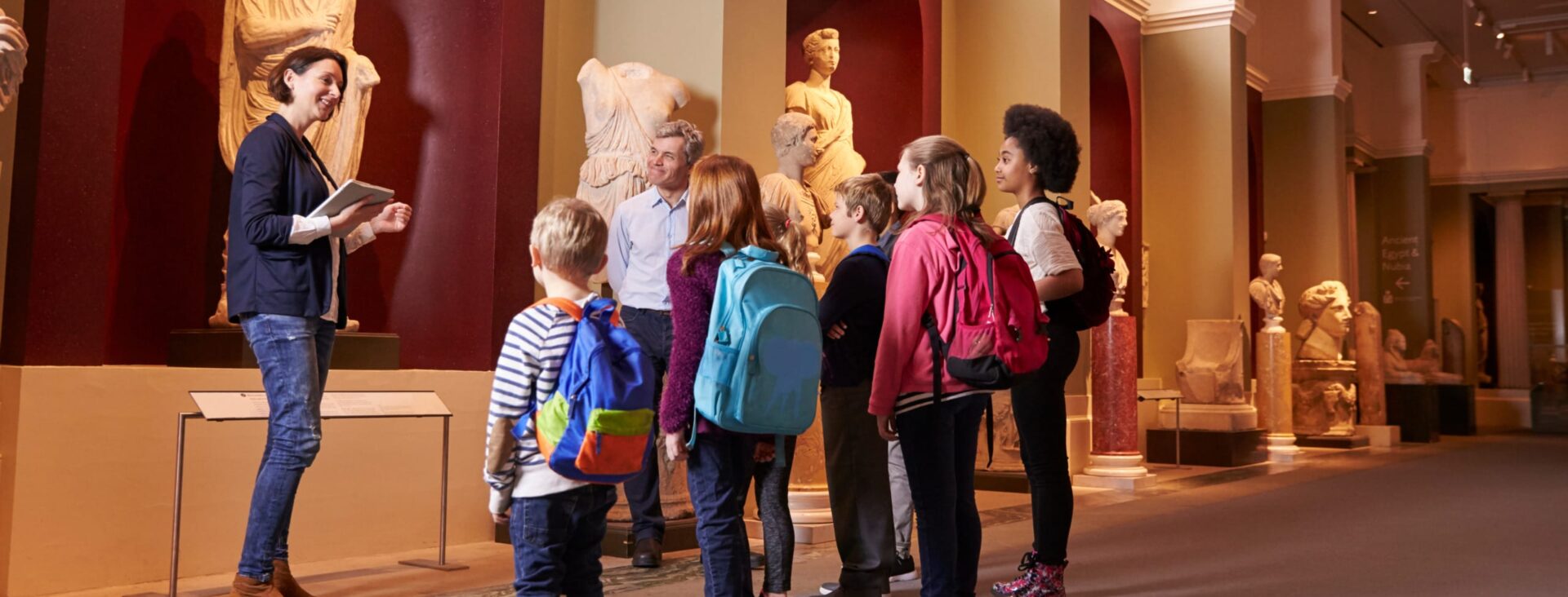 Museum guide leads a group of children on a tour, discussing sculptures in a gallery filled with classical art exhibits.