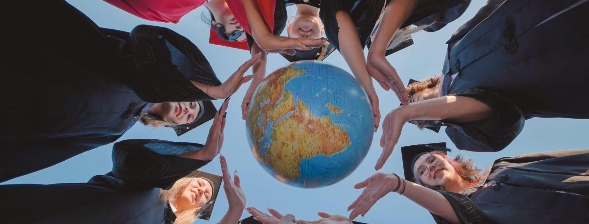 Graduates in caps and gowns standing in a circle holding a globe, viewed from below against a clear blue sky.