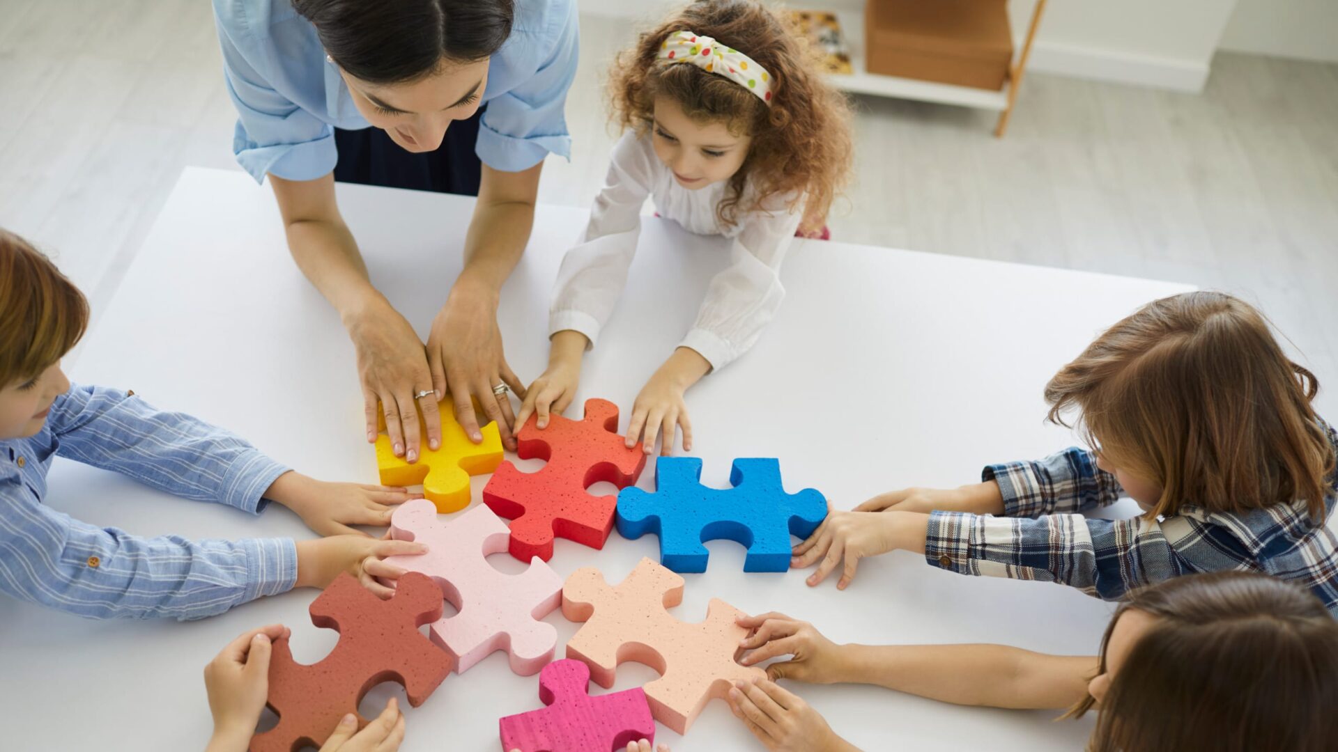 Children and a teacher collaborating to assemble large, colorful puzzle pieces on a white table.