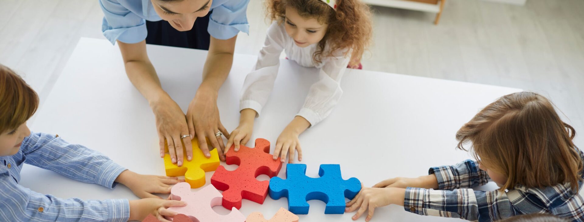 Children and a teacher collaborating to assemble large, colorful puzzle pieces on a white table.