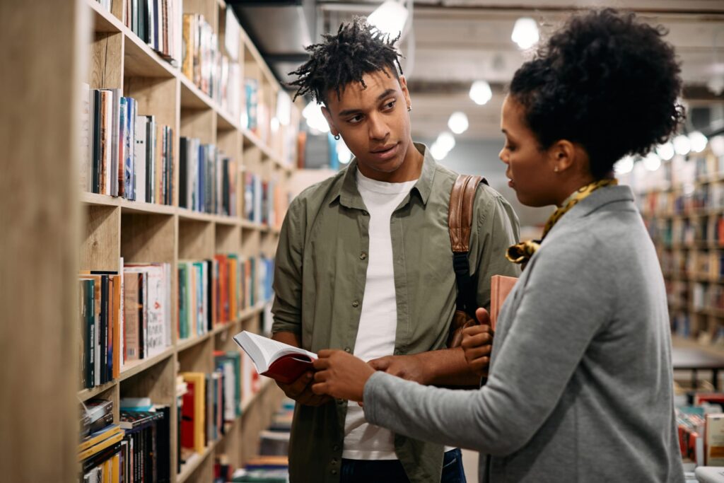 Two people discussing a book in a library aisle filled with shelves of books.