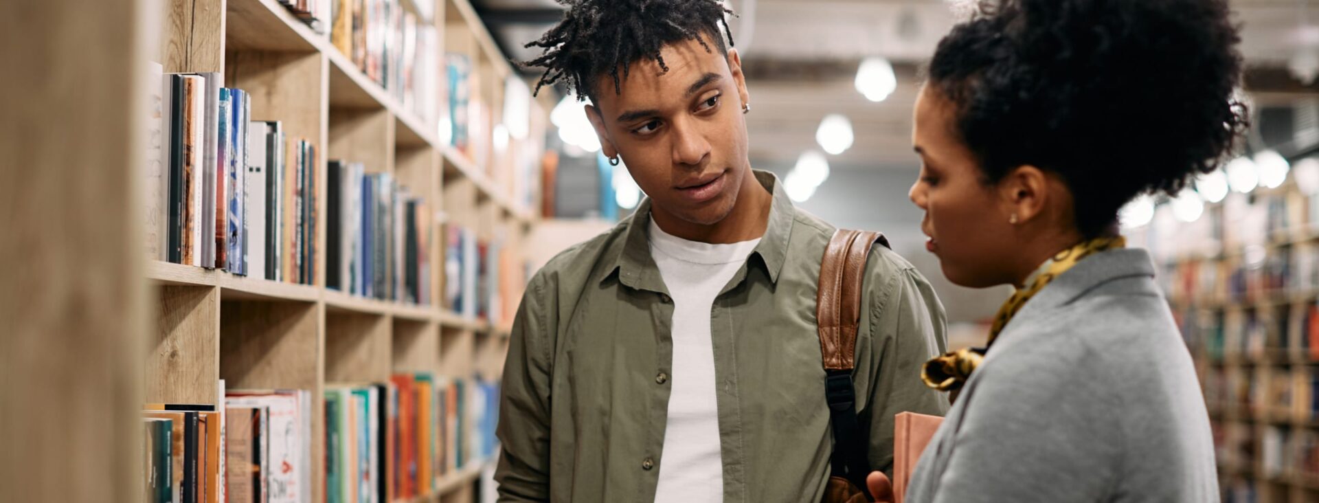 Two people discussing a book in a library aisle filled with shelves of books.