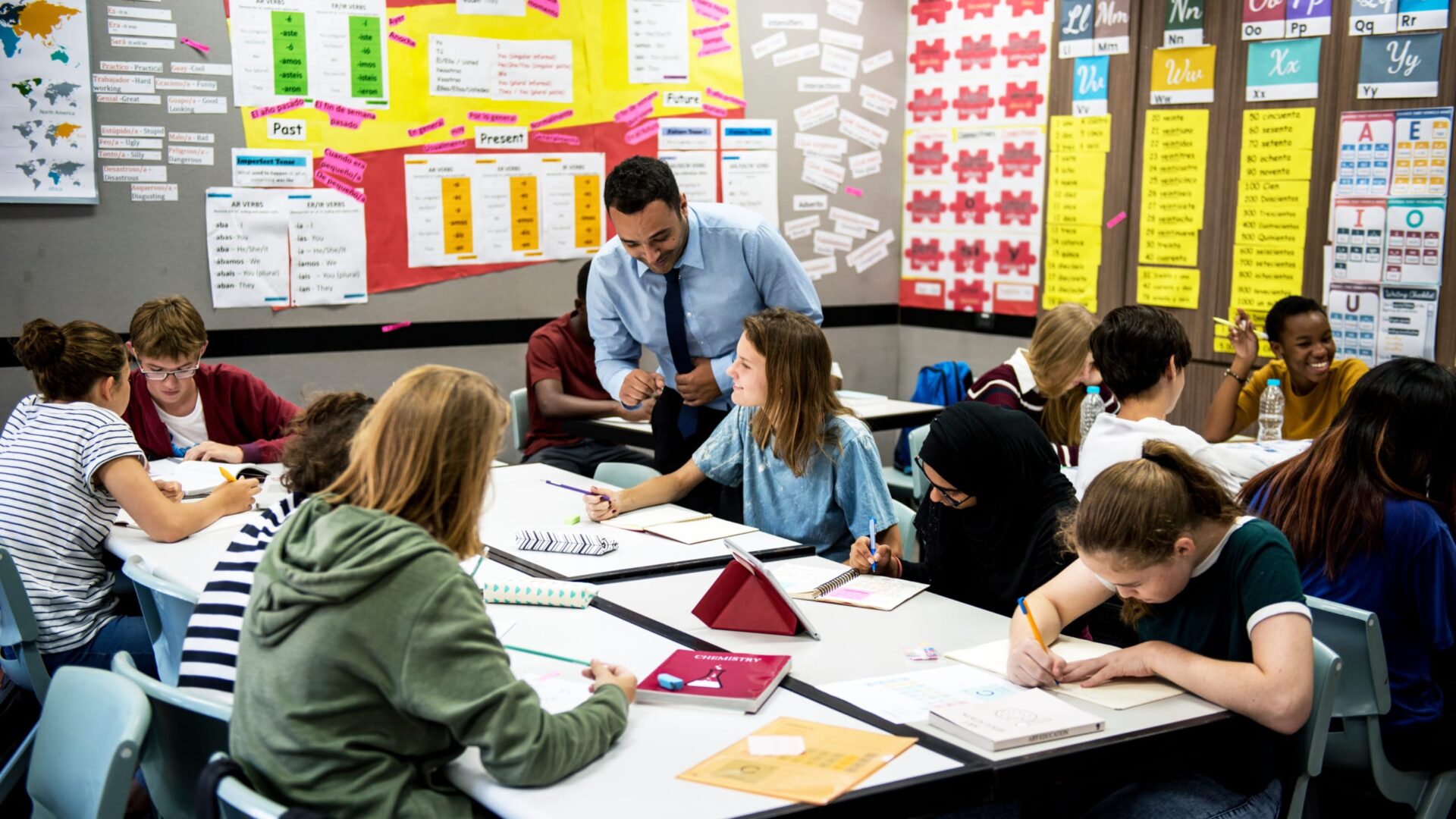 Teacher engaging with diverse students in a colorful classroom setting, with educational posters on the walls.