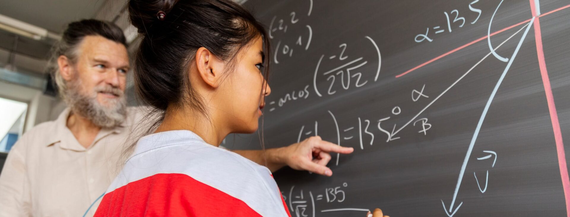 Student solving trigonometry on chalkboard with teacher's guidance in classroom setting.