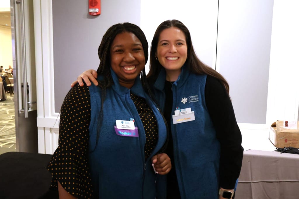 Two women smiling, wearing blue vests with name tags, standing indoors at an event.