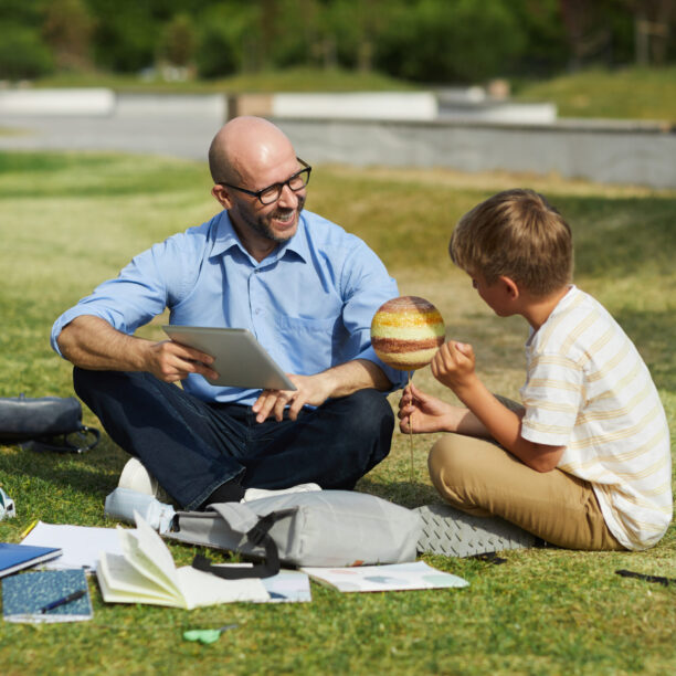 Man and boy sitting on grass discussing a model of planets with tablets and papers around them in a sunny outdoor setting.