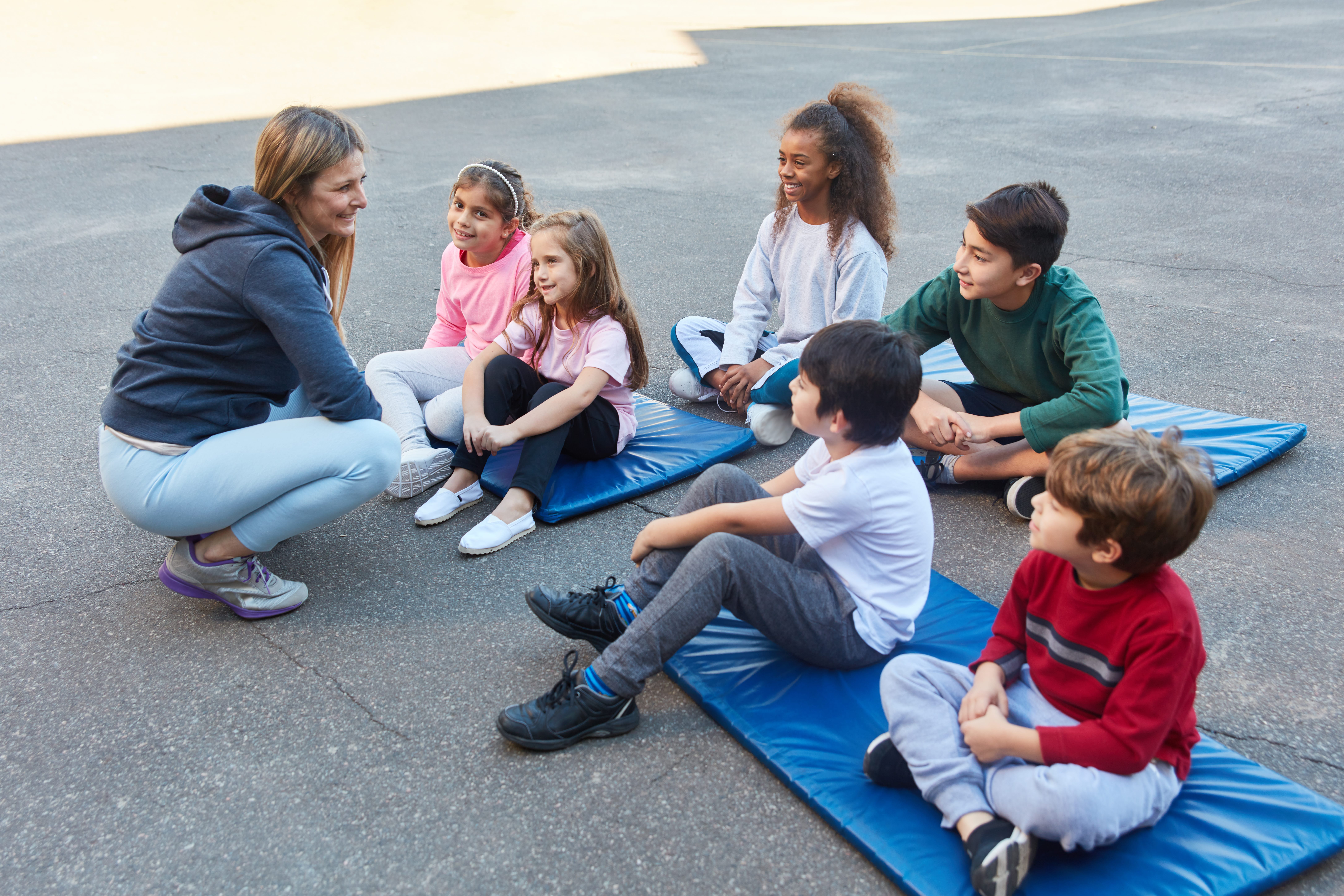 Instructor engaging with children seated on mats outside, participating in an outdoor activity session.