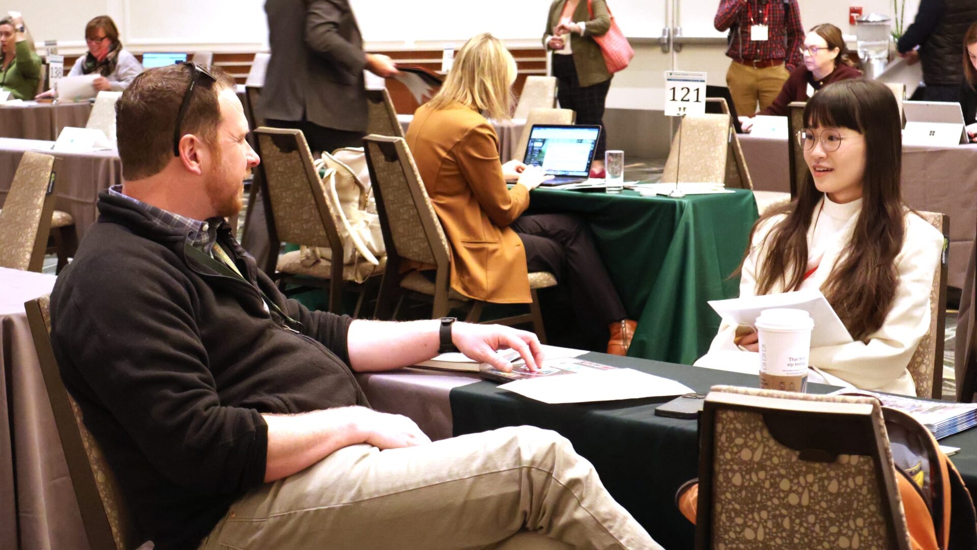 Two people having a discussion at a networking event with several others working in the background.