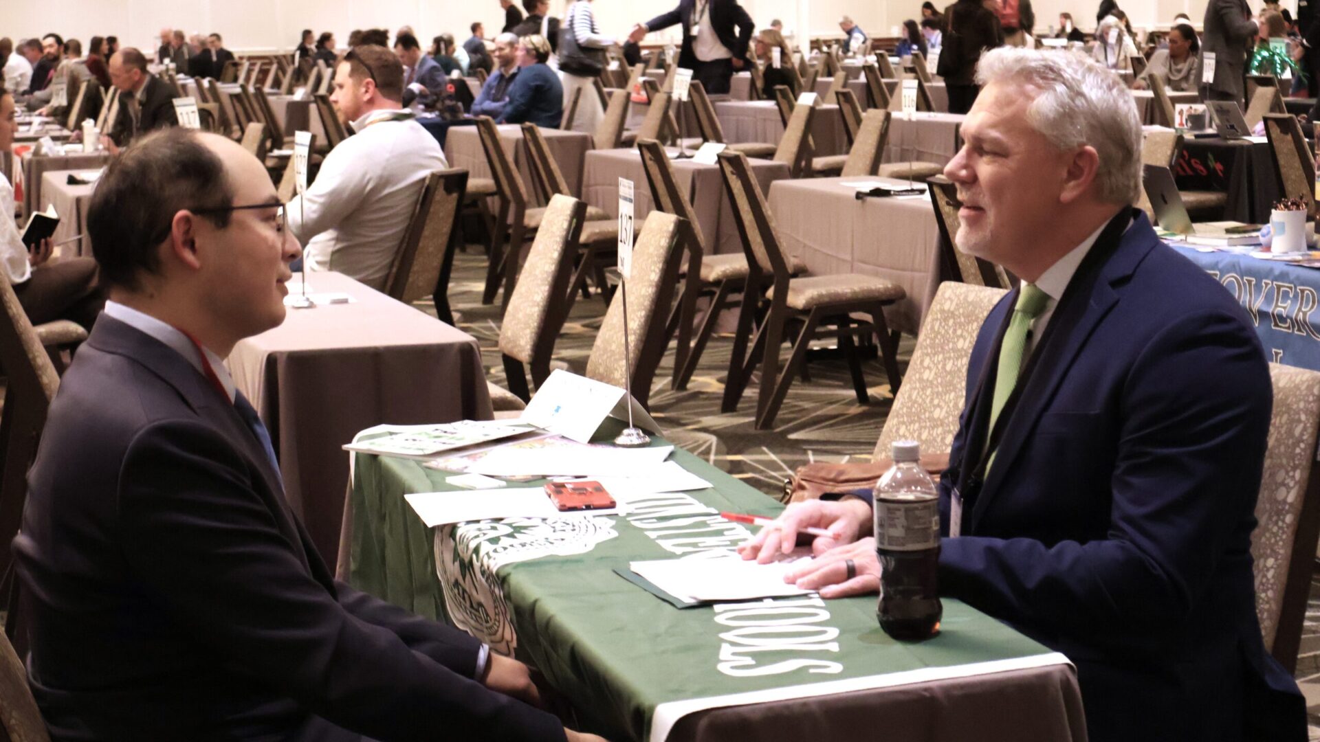 Two men in suits sitting at a table and talking, with people in the background at a professional event.