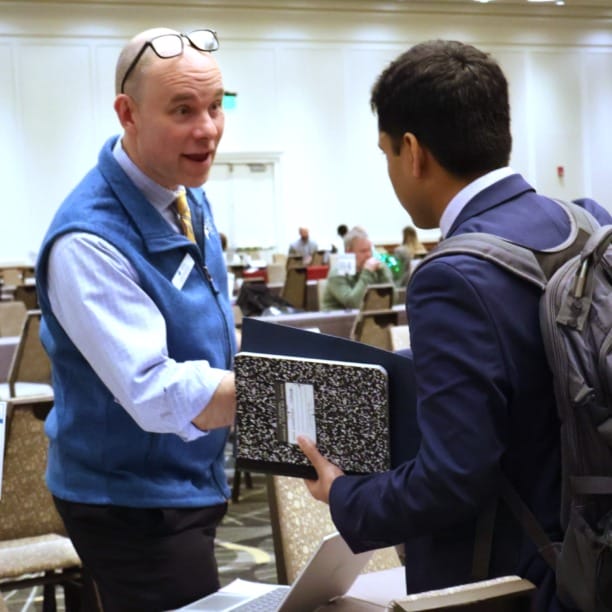 Business professional greeting a student with a handshake during a networking event in a conference room setting.