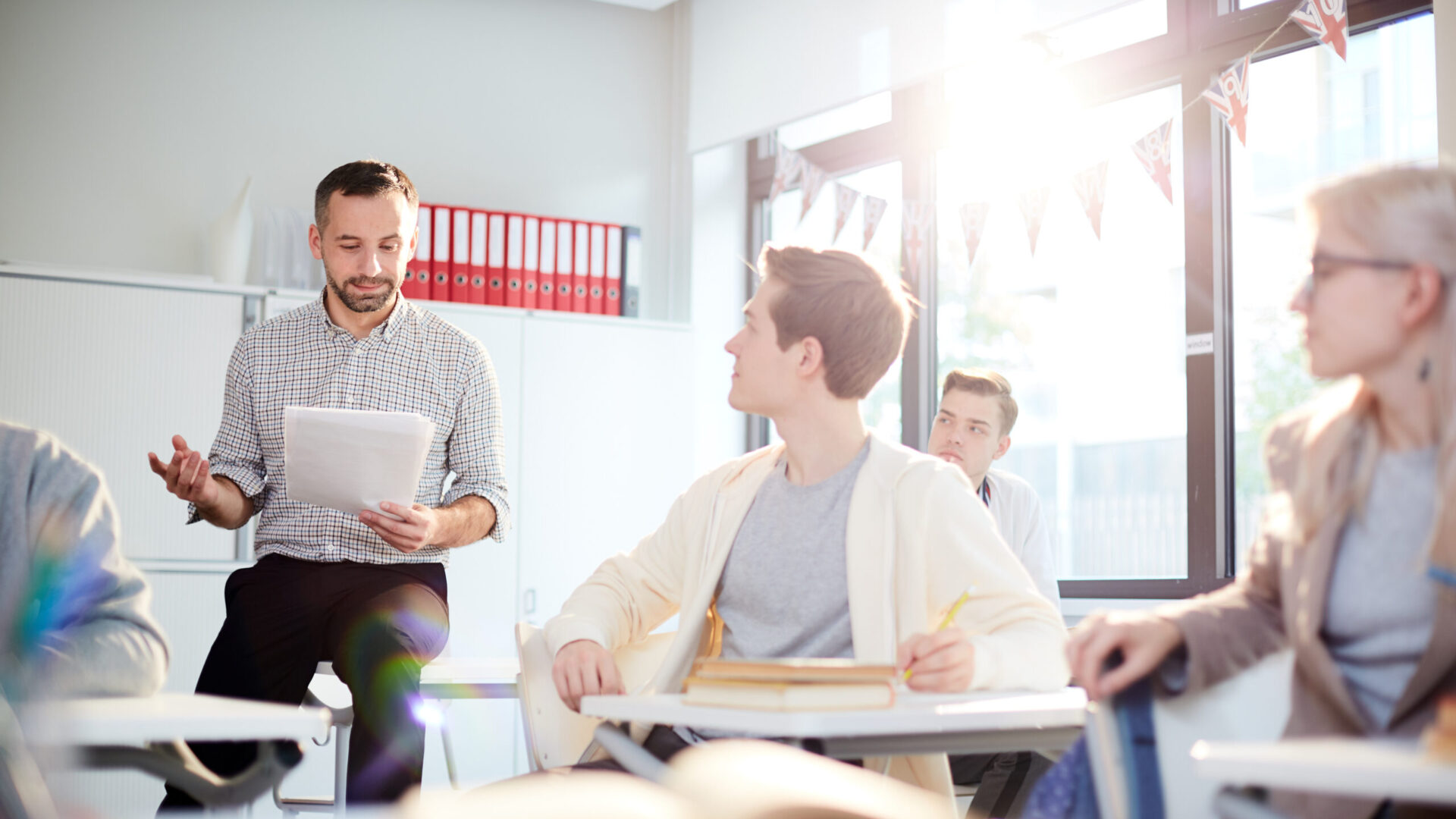 Teacher discussing with students in a classroom, sunlight streaming through the window, books and notes on desks.