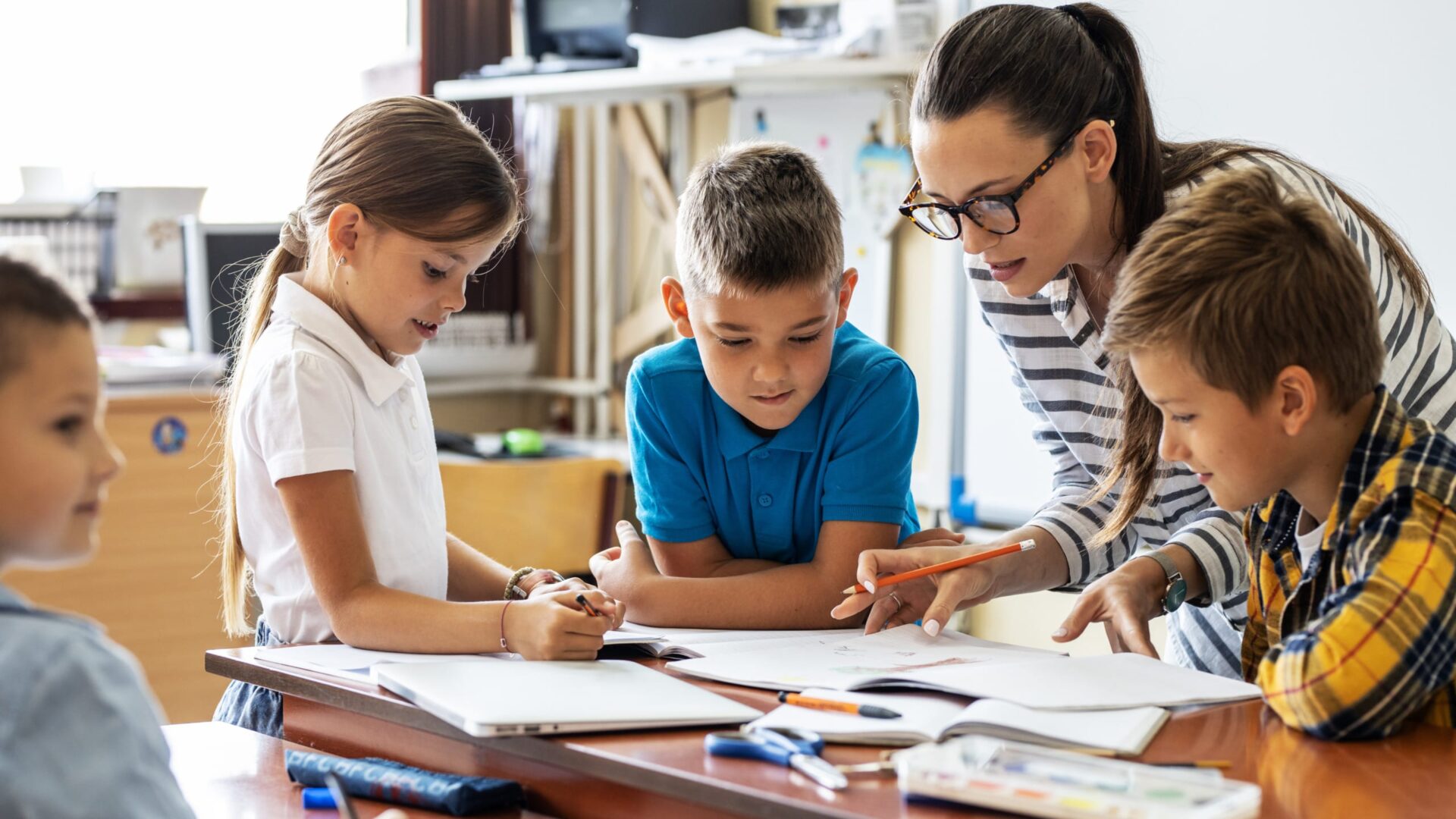 Teacher helping primary school students with classwork at a desk.