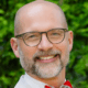 Smiling man with glasses and a red bow tie in front of green foliage background.