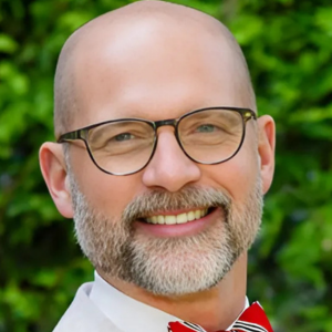 Smiling man with glasses and a red bow tie in front of green foliage background.