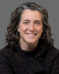 Smiling person with shoulder-length dark hair, wearing a textured black sweater and hoop earrings, against a gray background.