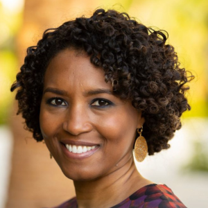 Smiling person with short curly hair and large earrings, standing outdoors with a blurred background.