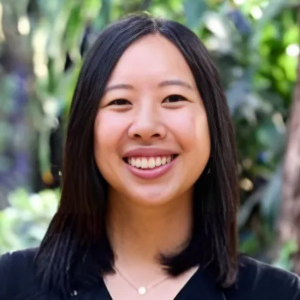 Smiling person with dark hair in a black shirt, standing in front of a blurred green leafy background.