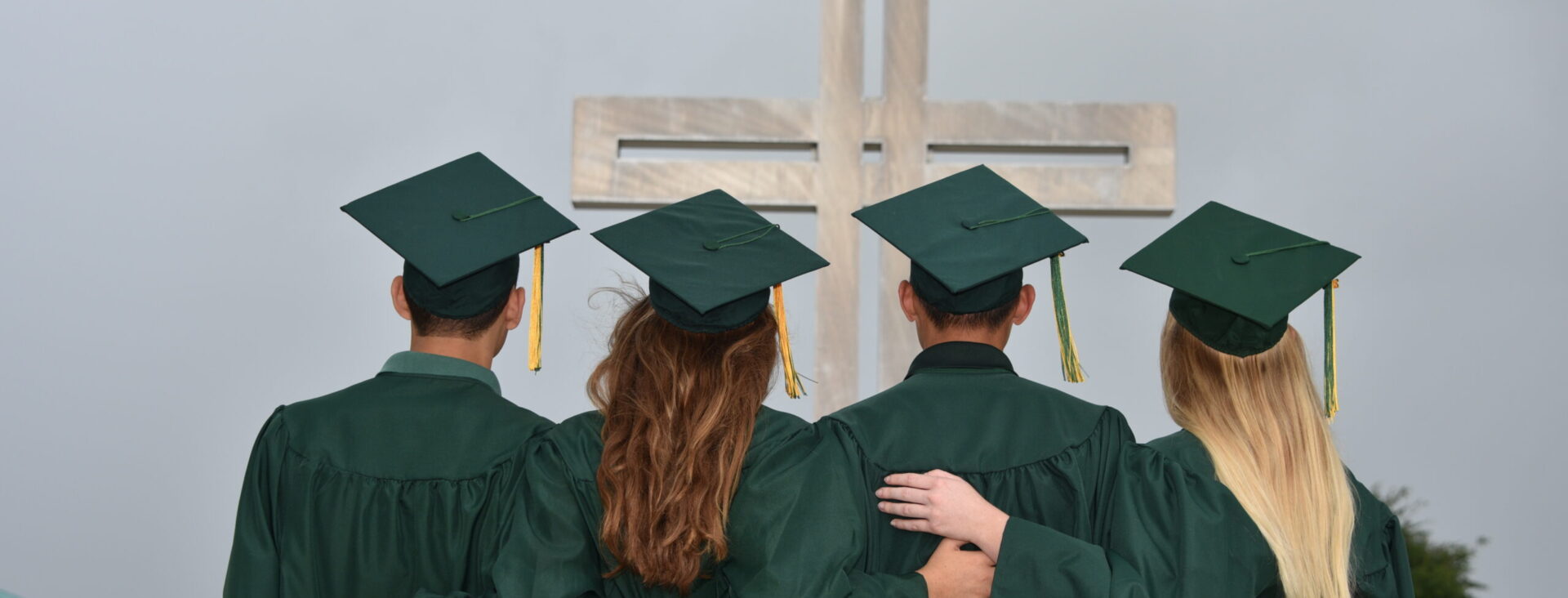 Graduates in green gowns and caps standing with arms linked, facing a large cross in the background.