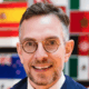 Man with glasses smiling, wearing a suit, international flags in the background