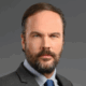 Man with a beard wearing a suit and tie against a gray background