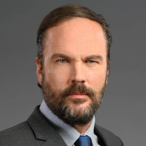 Man with a beard wearing a suit and tie against a gray background