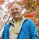 Man in a blue jacket smiling outdoors with colorful autumn leaves in the background.