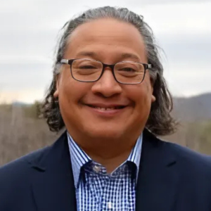 Smiling person with glasses wearing a blue blazer and checkered shirt, standing outdoors with trees and a cloudy sky in background.