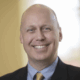 Smiling bald man in a suit and tie, looking directly at the camera in a warmly lit room.