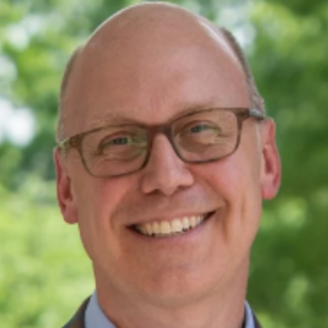 Smiling bald man with glasses in a suit, standing outside with a blurred green background.