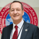 Man in a suit and red tie standing in front of a round sign with text, smiling at the camera.