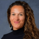 Smiling person with long curly hair and hoop earrings wearing a black turtleneck against a gray background.