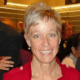 Person in a red jacket smiling at an indoor event with people in the background.