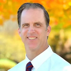 Man smiling outdoors with autumn leaves in the background, wearing a white shirt and striped tie.